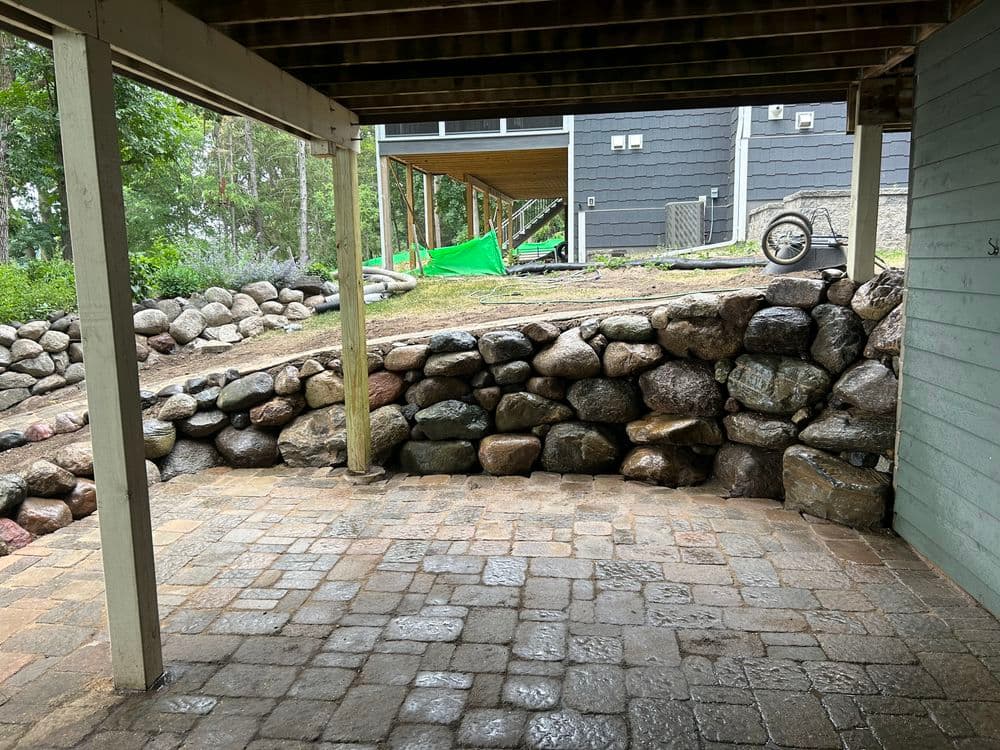 Patio with stone wall and greenery, featuring a rustic design under a wooden structure.