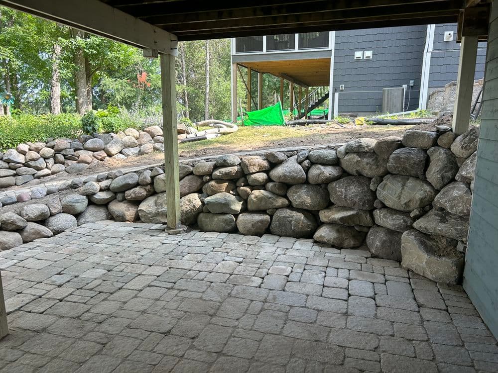 Stone wall and paved patio area beneath a raised deck, surrounded by greenery and construction materials.