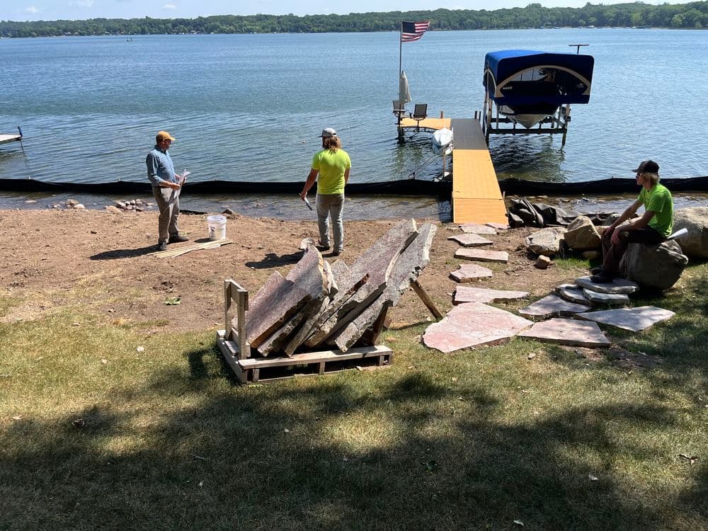 Workers installing stone pathways by a lake with a boat dock and American flag backdrop.