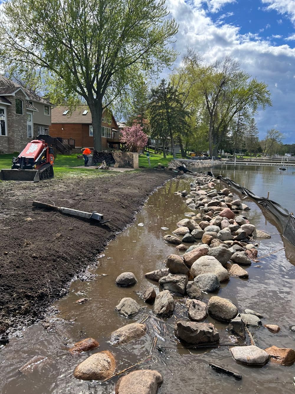 Excavator working on beach restoration with rocks along a lakefront under a blue sky.