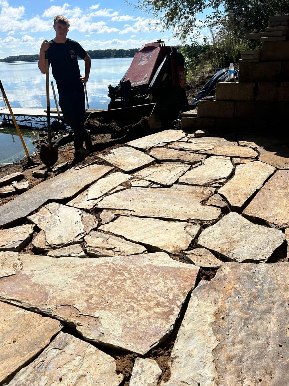 Person working on a stone pathway by a lake, with landscaping equipment in the background.