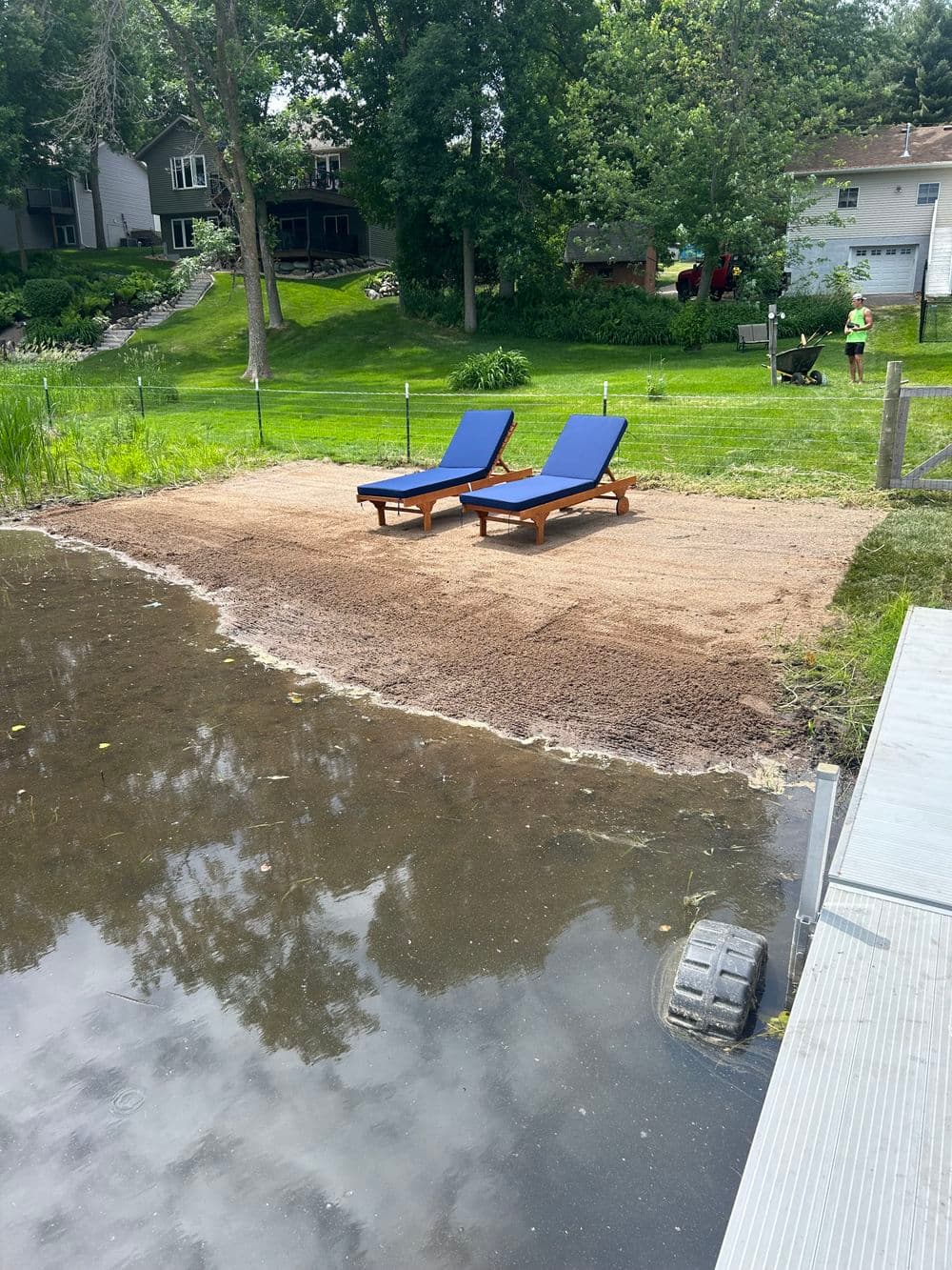 Lounge chairs on a sandy lakeside patio with green trees and a house in the background.