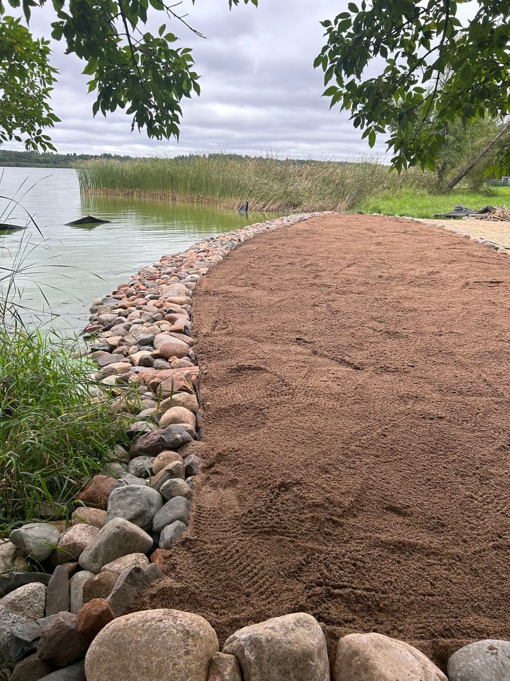 Beach shoreline with sandy area bordered by smooth stones and lush green grass.