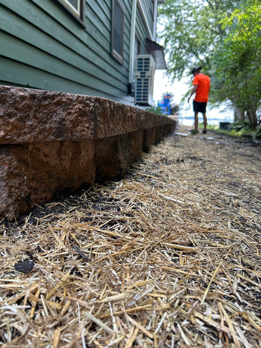 Pathway beside a house with a stone border and straw ground cover, person walking in background.