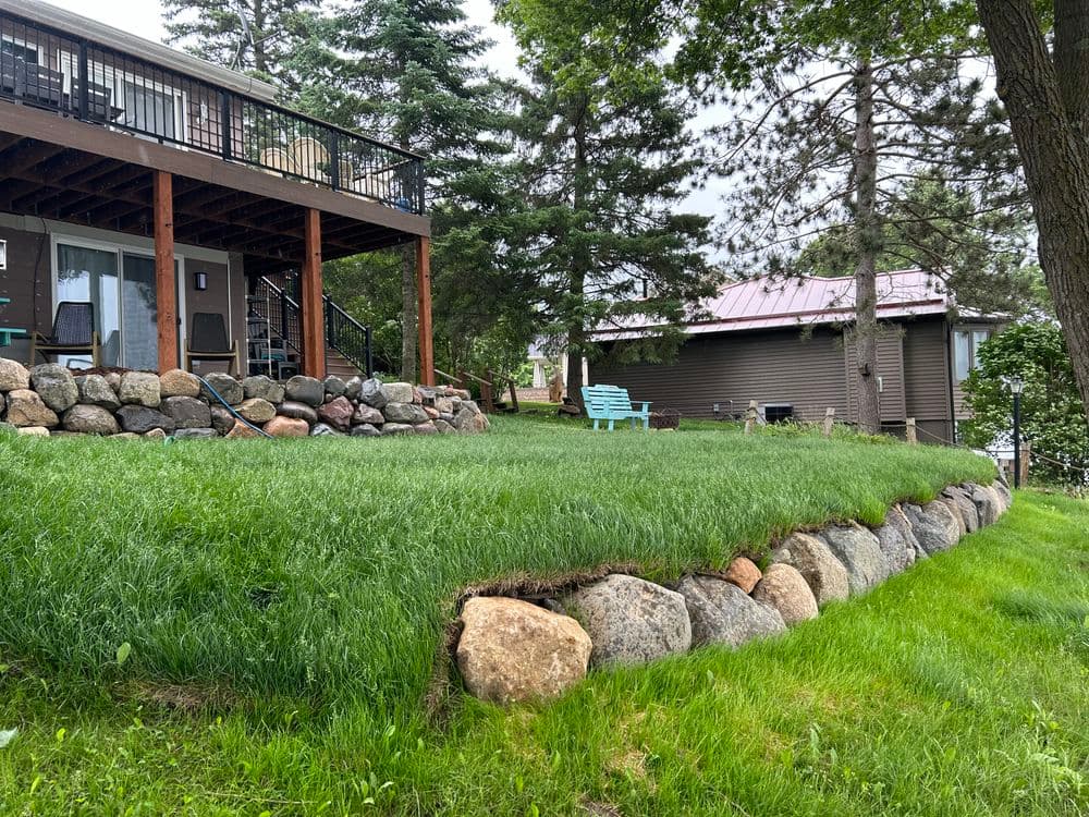 Charming lakeside property with a green lawn, stone border, and wooden deck surrounded by trees.