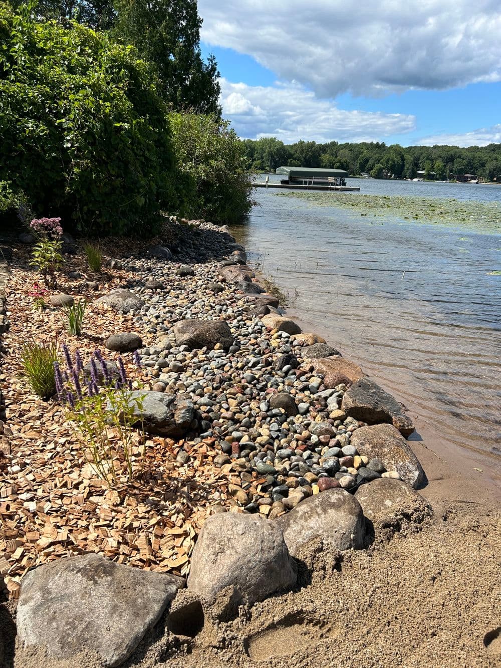 Lake shoreline with pebbles, plants, and a boat dock under a partly cloudy sky.