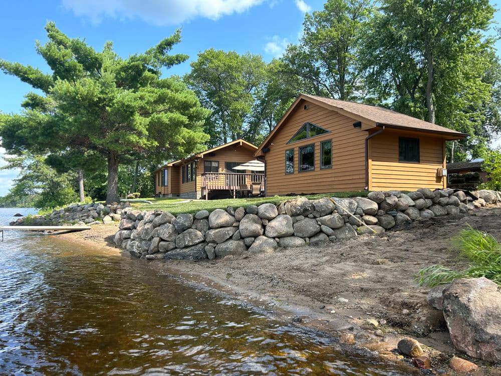 Scenic lakeside cabins with stone shoreline and lush greenery on a sunny day.