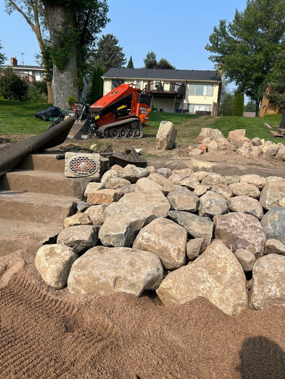 Construction site with rocks, sand, and a skid steer loader in a residential yard.