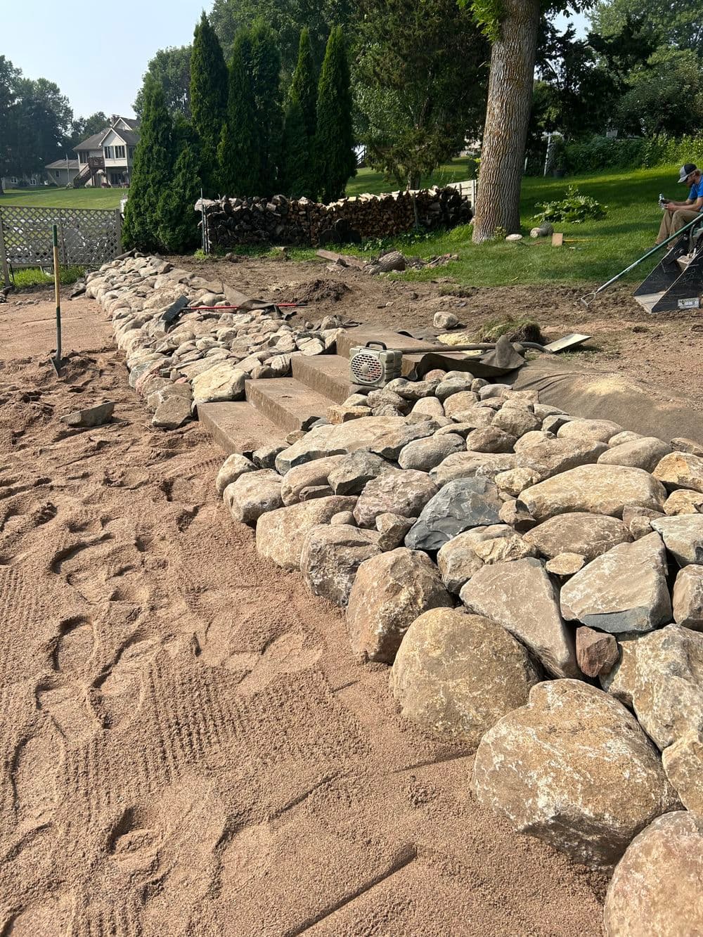 Landscape featuring a stone wall under construction, surrounded by sand and greenery.