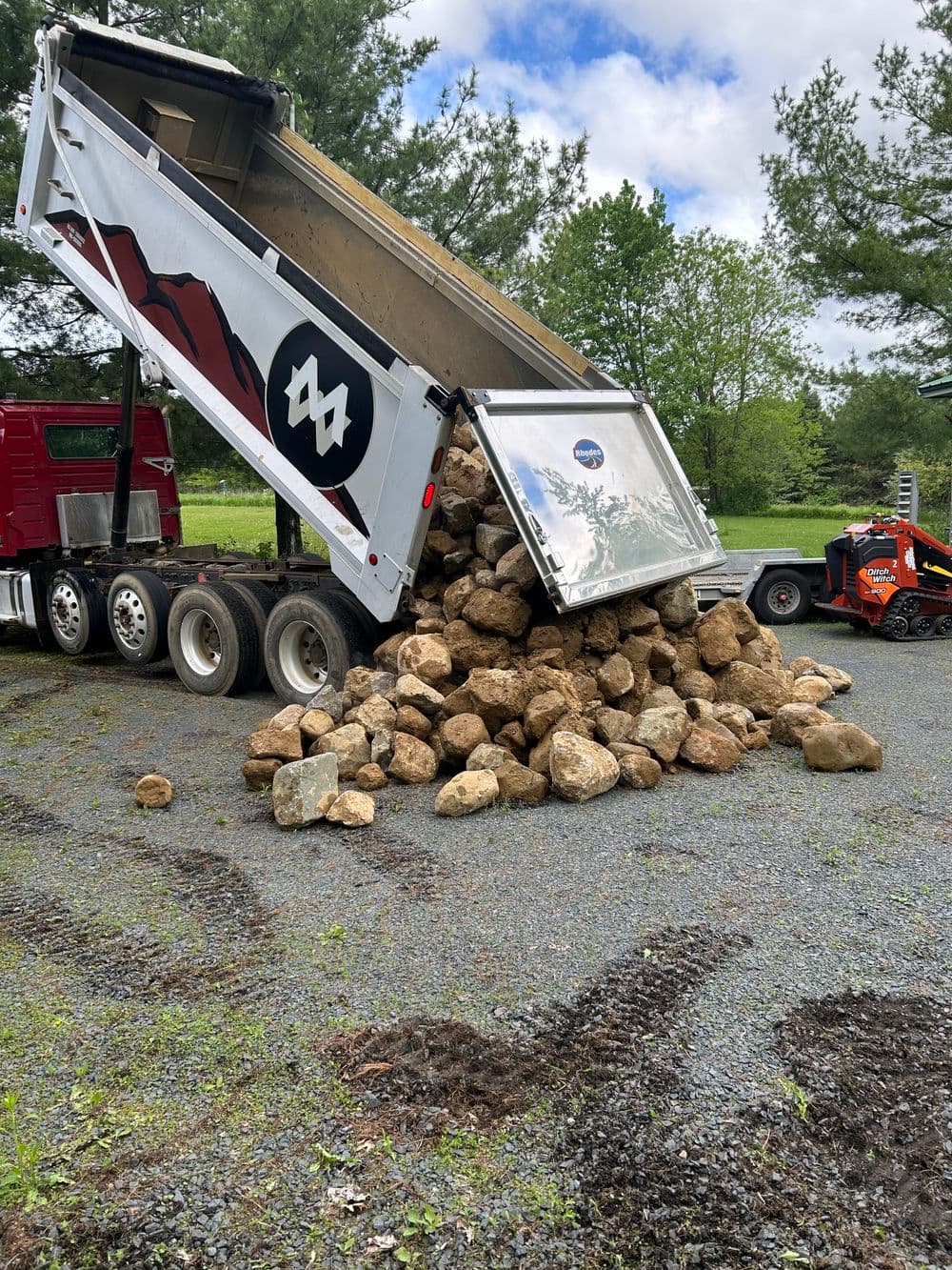 Dump truck unloading large rocks onto gravel surface in a wooded area.