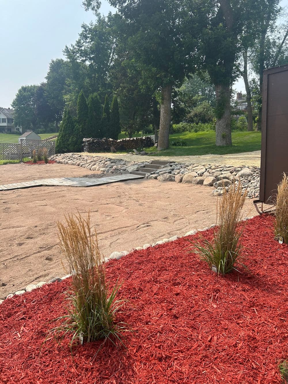 Landscape with red mulch, ornamental grasses, and a rocky path in a sunny backyard setting.