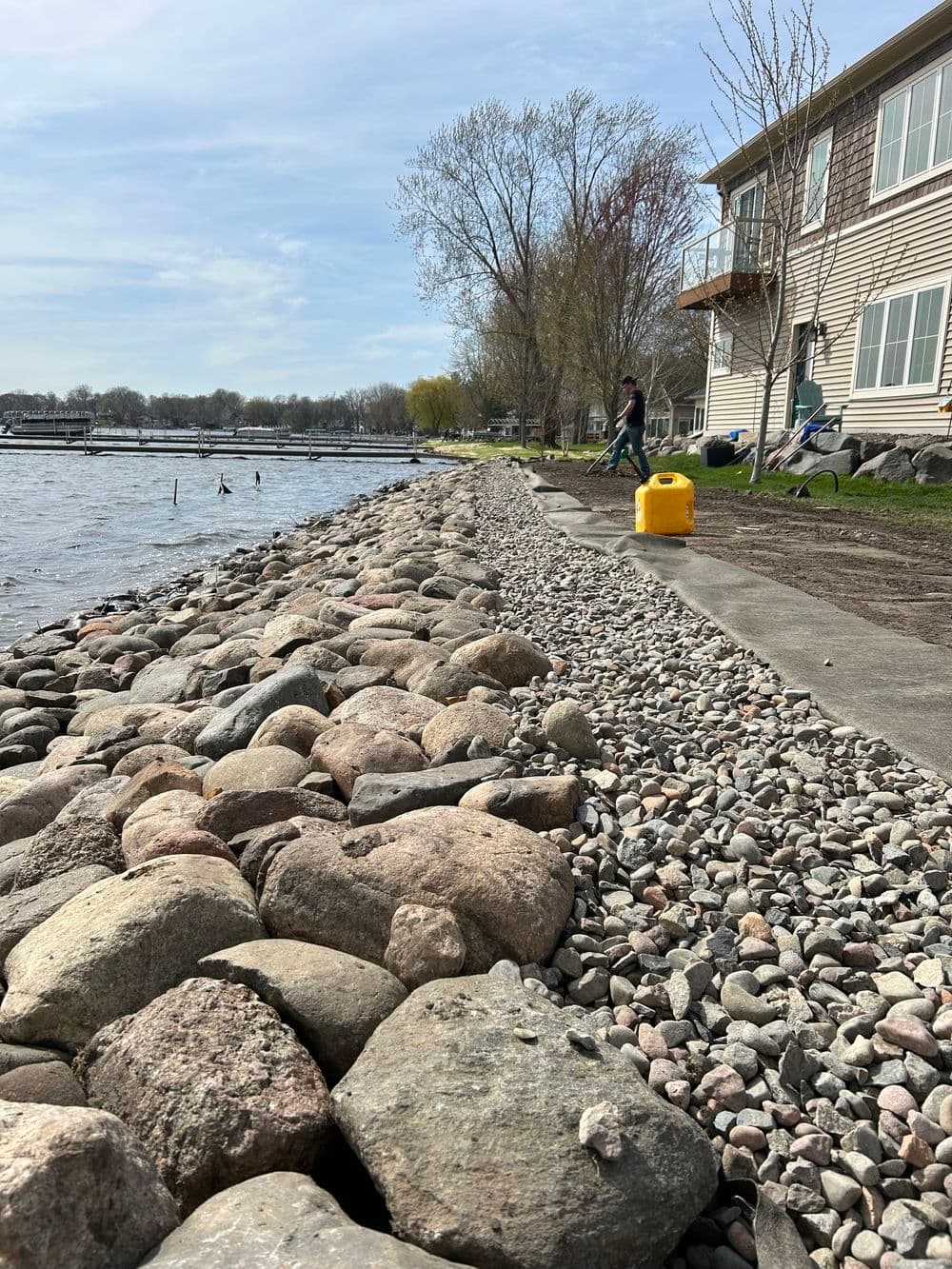 Lakeside shoreline with rocks and gravel near a building, featuring a person and water.
