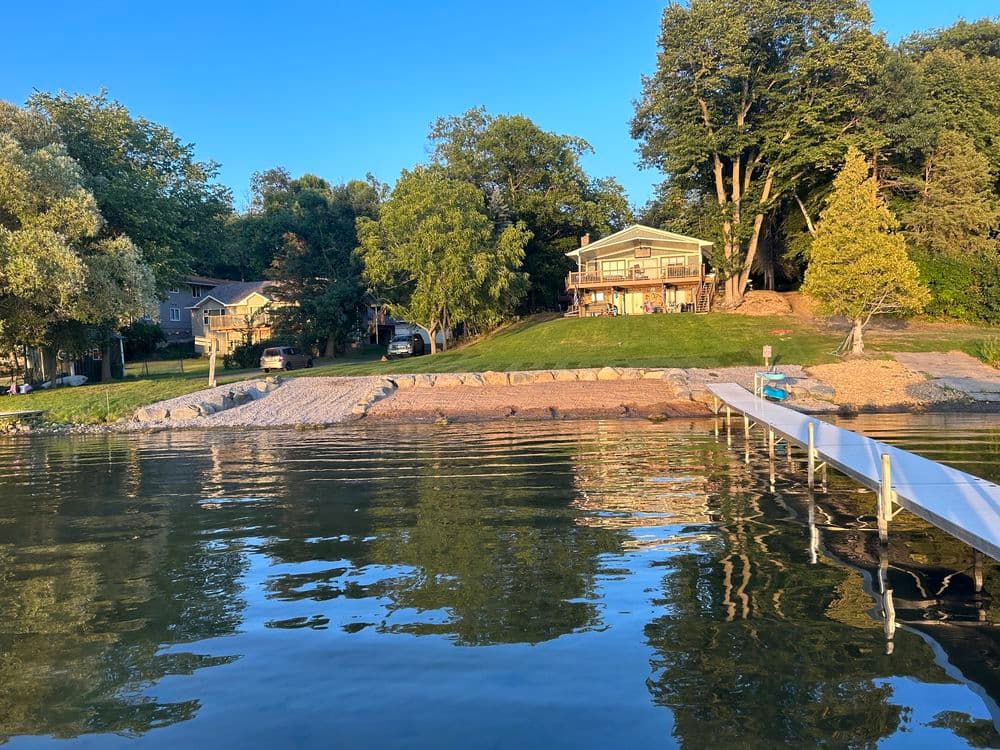 Lakefront property with a house, dock, and lush greenery on a sunny day.